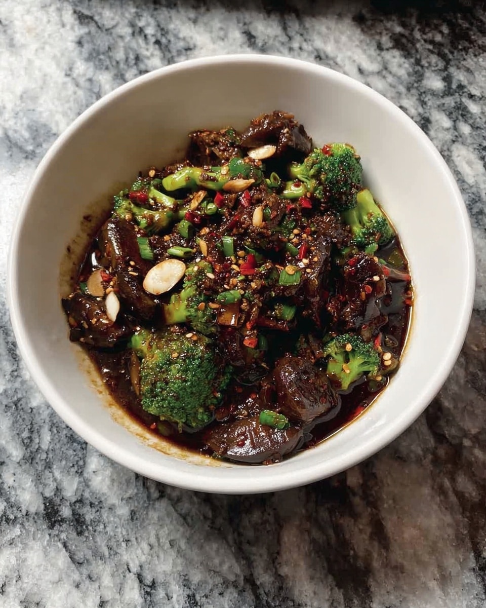 The image shows a white bowl filled with a dark, glossy stir-fry dish. The dish has several layers, with dark brown caramelized pieces of meat or mushrooms on top, mixed with small green broccoli florets and sprinkled with light-colored sliced garlic and chili flakes for texture. The sauce covers the ingredients well, giving a shiny look. The bowl is sitting on a white marbled surface. Photo taken with an iphone --ar 4:5 --v 7