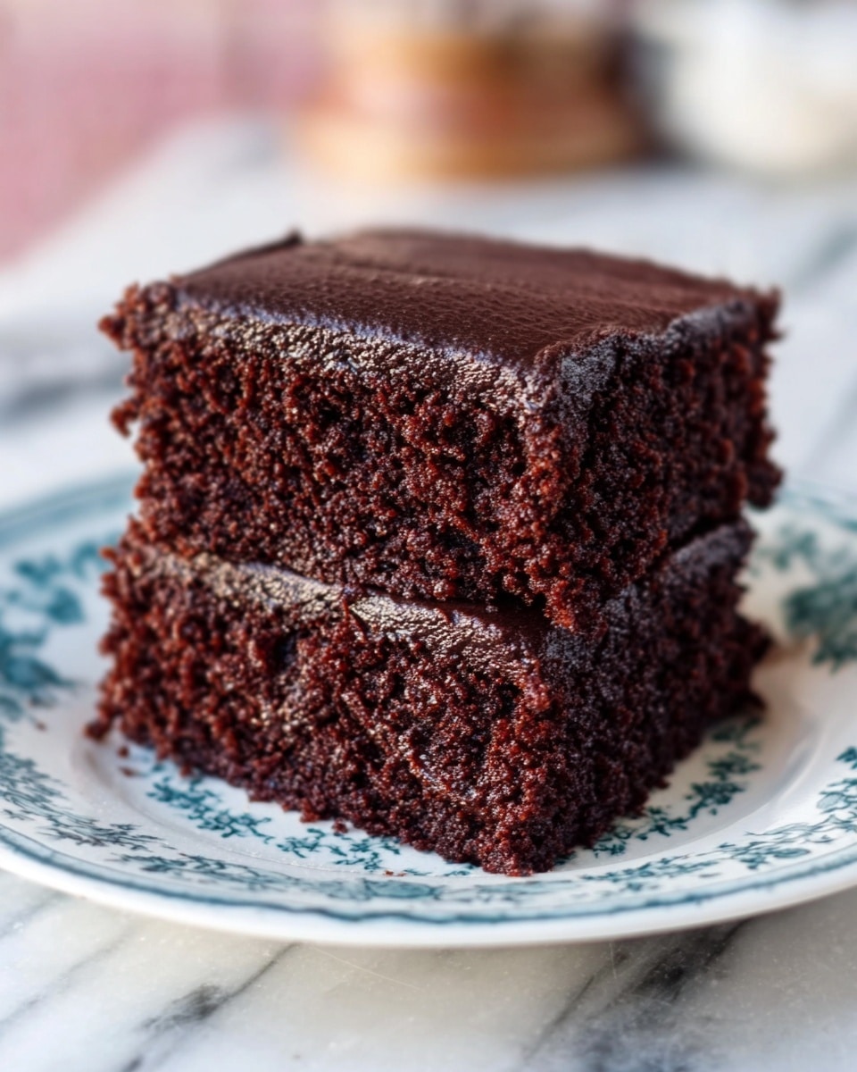 The image shows two pieces of rich, dark chocolate cake stacked on top of each other on a white plate with blue floral patterns. The cake layers are thick and moist with a slightly crumbly texture visible on the sides. The top layer has a smooth, shiny chocolate frosting covering it completely. The plate sits on a white marbled surface with a soft focus background showing some blurred kitchen items. photo taken with an iphone --ar 4:5 --v 7