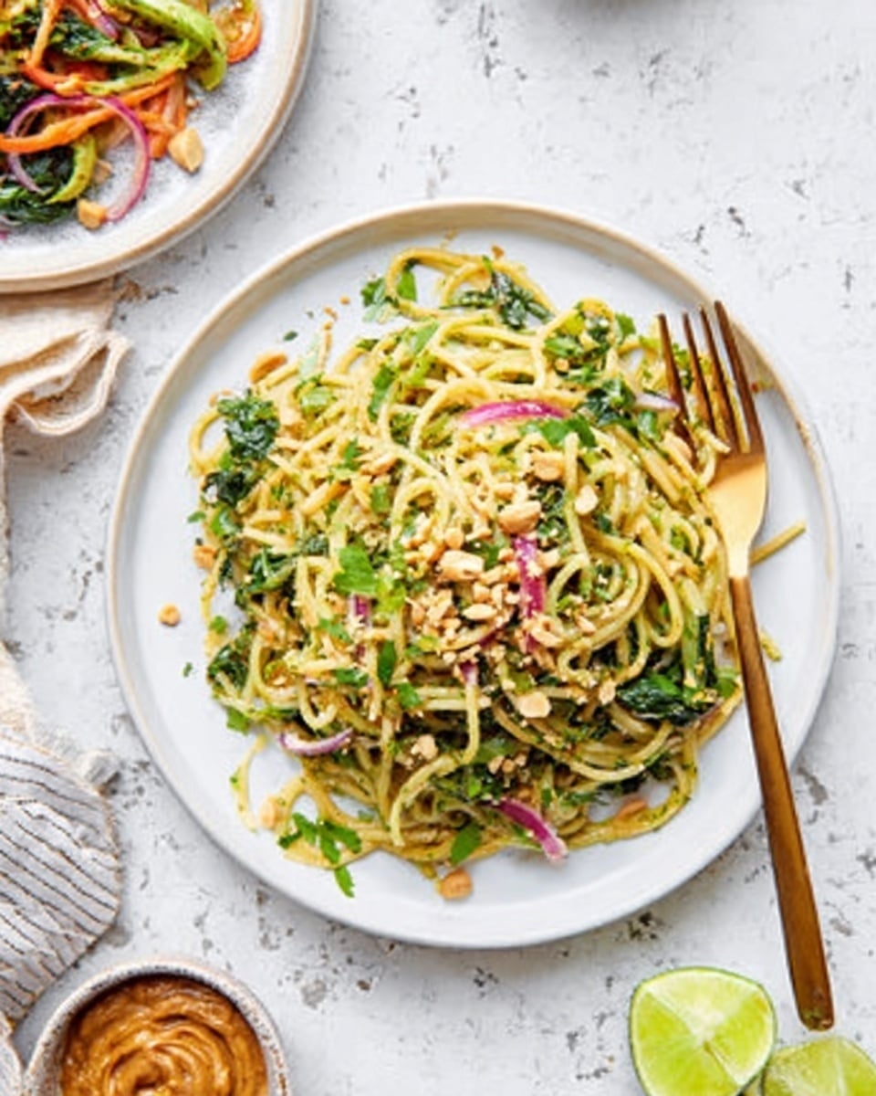 A white plate with a nest of thin noodles mixed with green leafy vegetables, red onion slices, and bits of crushed peanuts on top. The noodles look soft and shiny, with a light green sauce coating them evenly. There are fresh herbs sprinkled over, adding color contrast. A golden fork rests on the plate’s right side, while a white marbled surface serves as the background. In the top left corner, a partial view of another white plate with more noodles and vegetables is visible, along with a small bowl of brown sauce or paste nearby. A wedge of lime is at the bottom right of the scene, adding fresh green color. photo taken with an iphone --ar 4:5 --v 7