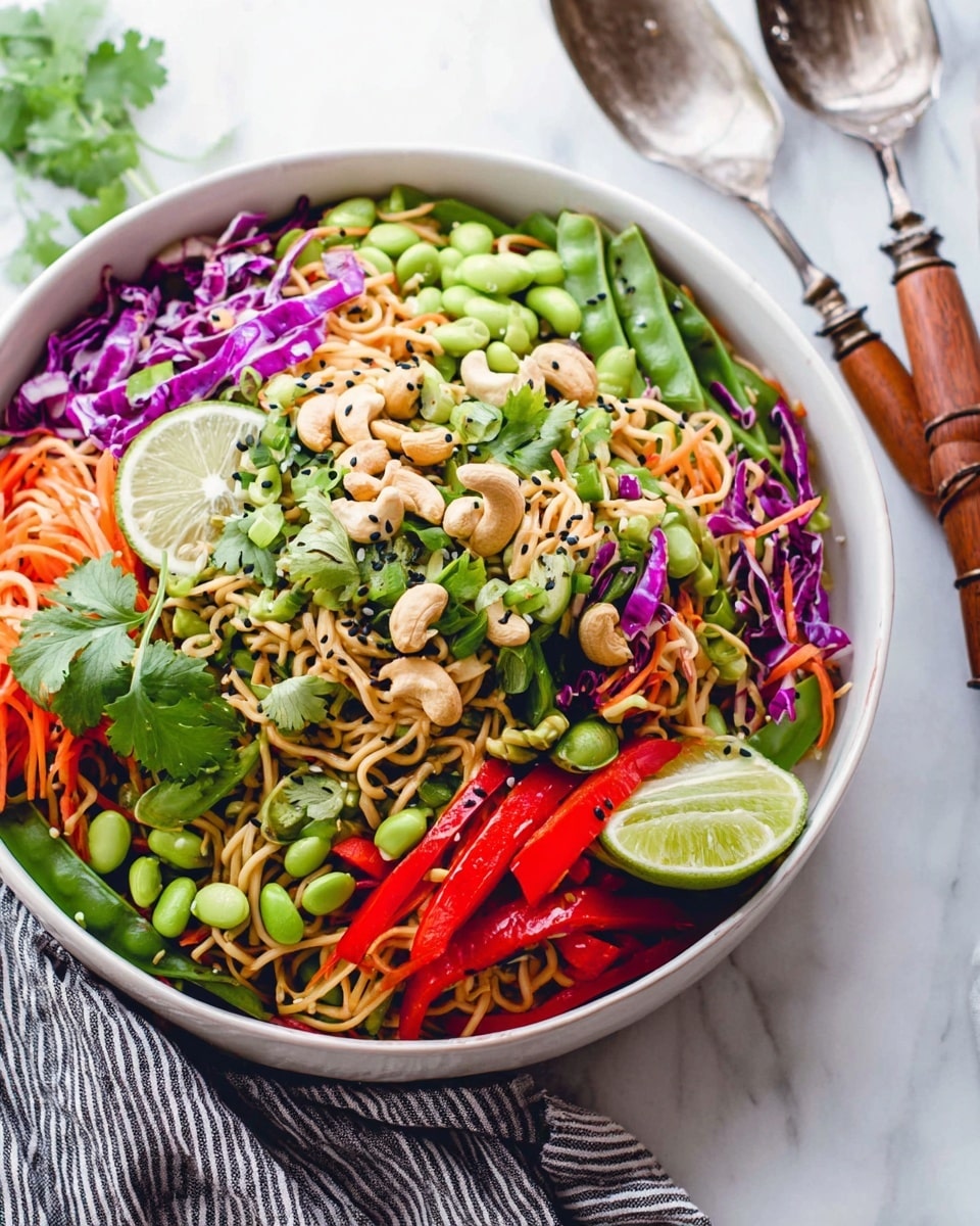 A large white bowl filled with a colorful noodle salad sits on a white marbled surface with a striped cloth nearby holding a fork and spoon with wooden handles. The salad has three main layers: at the bottom are curly, light brown noodles; the middle layer has sliced red bell peppers, shredded purple cabbage, and green snap peas; the top layer is garnished with whole cashews, chopped green onions, light green edamame beans, fresh cilantro leaves, sesame seeds, and a half lime sprinkled with black sesame seeds. Photo taken with an iphone --ar 4:5 --v 7