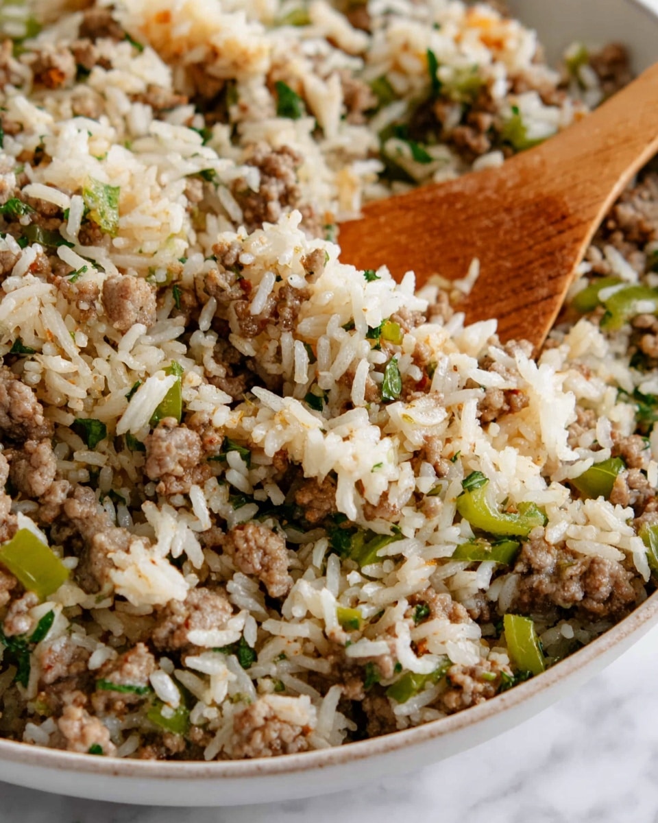 The image shows a close-up of a white bowl filled with a mixture of cooked white rice, browned ground meat, and small green bell pepper pieces evenly mixed throughout. The texture is loose and slightly oily, with bits of cooked onion and small herbs scattered within. A wooden spoon lifts a scoop of the mixture, highlighting the moist, tender grains of rice and finely crumbled meat combined with the vibrant green pepper chunks against the white interior of the bowl. The surface underneath the bowl has a white marbled texture. photo taken with an iphone --ar 4:5 --v 7
