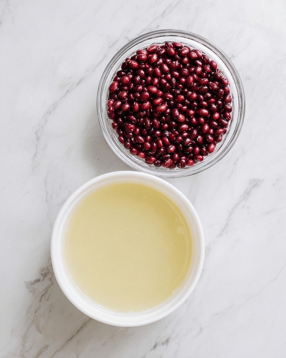 The image shows two bowls on a white marbled surface, positioned diagonally. At the top is a clear glass bowl filled with small, shiny, dark red beans with a smooth texture. At the bottom is a white bowl with a pale yellow liquid that looks slightly thick and smooth. The bowls are clean and simple in design, and the scene is bright with natural light. photo taken with an iphone --ar 4:5 --v 7