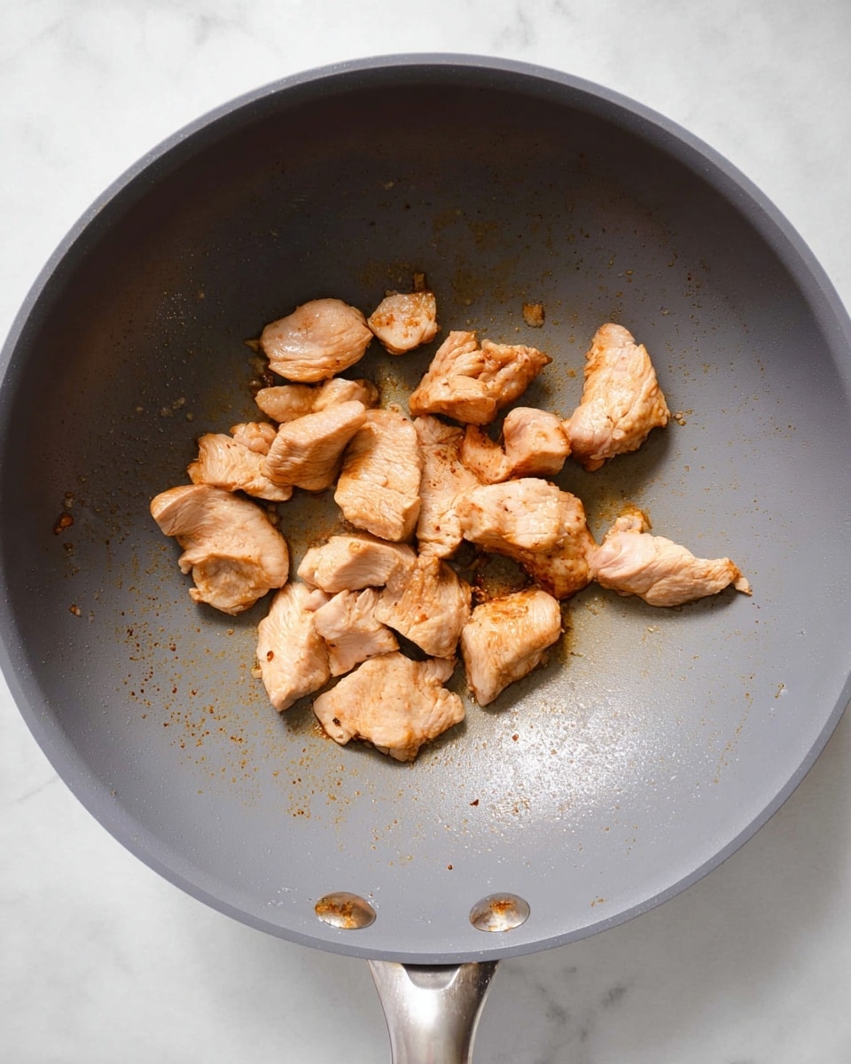 Inside a grey non-stick frying pan placed on a white marbled surface, there are several pieces of cooked light brown meat scattered slightly apart from each other. The meat pieces have a soft, uneven texture with some areas showing a slight golden brown sear while others are lighter. The pan’s surface is smooth and clean except for some brown spots where the meat has touched it. The handle of the pan extends to the lower right side. Photo taken with an iphone --ar 4:5 --v 7