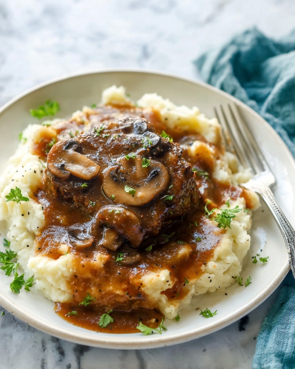 A white plate holds a base layer of creamy mashed potatoes with small green herb pieces mixed in, showing a soft and slightly textured surface. On top is a thick brown piece of meat smothered in rich dark brown mushroom gravy, with visible small mushroom slices and specks of black pepper. Fresh green parsley leaves are scattered over the dish for garnish. The plate sits on a white marbled surface with a silver fork at the edge. photo taken with an iphone --ar 4:5 --v 7