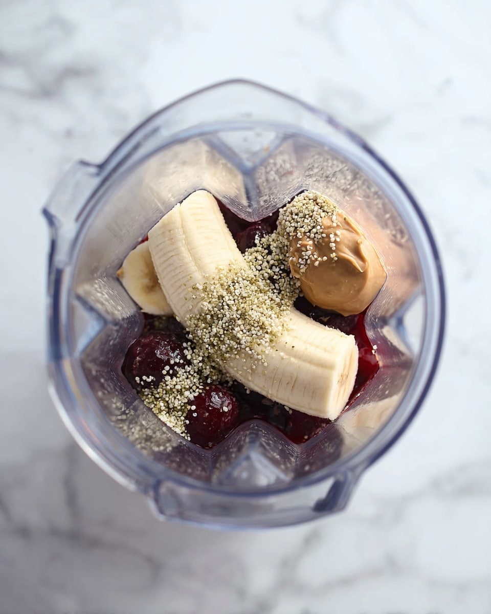 Inside a clear blender, there is one half banana placed diagonally in the middle, surrounded by dark red frozen cherries at the bottom layer. A small scoop of light brown peanut butter rests near the banana on one side. On top of the banana and cherries, there is a sprinkling of tiny beige seed-like bits, likely hemp seeds. The blender is set on a white marbled surface. The photo was taken with an iphone --ar 4:5 --v 7