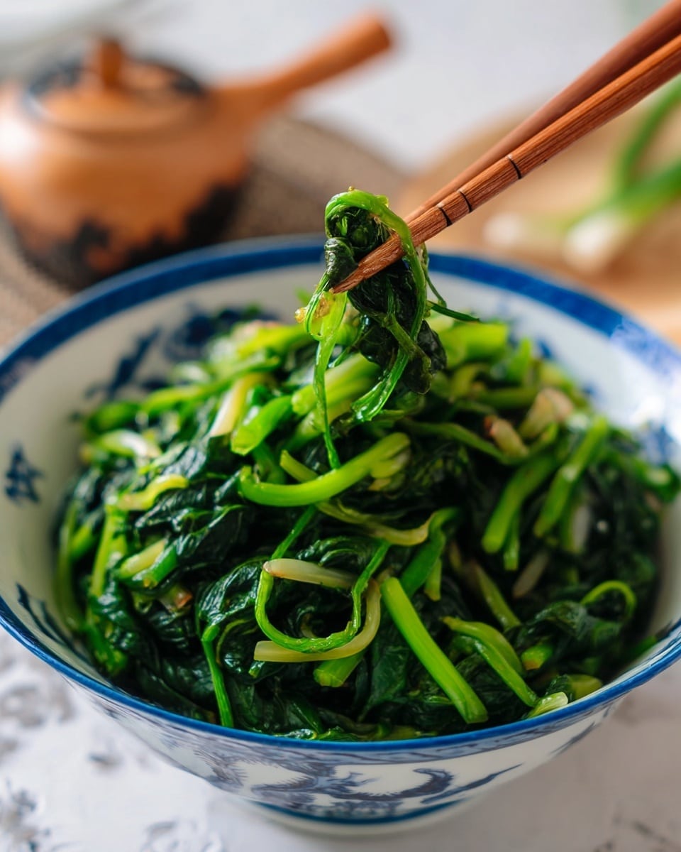 A close-up view of a white bowl with blue designs filled with cooked green leafy vegetables with long stems, showing a shiny and slightly wet texture. A pair of wooden chopsticks held by a woman's hand lifts a small bundle of the greens above the bowl, focusing on the deep green leaves and lighter green stems. The background features a white marbled texture and some blurred items, keeping the focus on the vibrant greens in the bowl. photo taken with an iphone --ar 4:5 --v 7