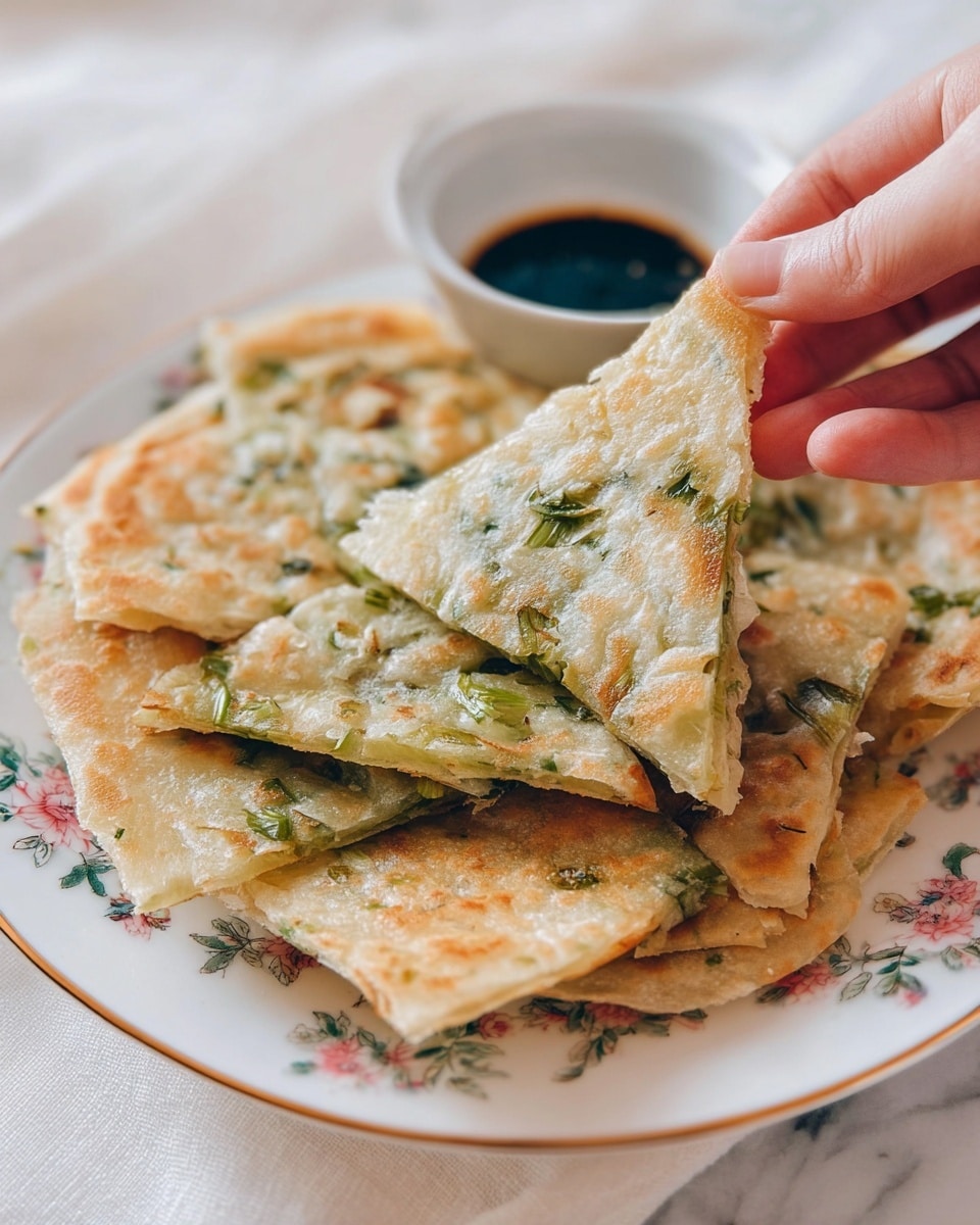 A close-up of a white plate with floral patterns holds several pieces of thin, crispy scallion pancakes stacked unevenly. The pancakes have a light golden brown color with green scallion pieces spread throughout each layer, giving a textured look. A woman’s hand is lifting one triangular piece, showing its light, flaky texture and the scallions embedded inside. In the background, a small white bowl filled with dark soy sauce adds contrast. The scene is set on a white marbled surface with soft natural light coming from the side, highlighting the pancakes’ crisp edges and slight bubbles. photo taken with an iphone --ar 4:5 --v 7