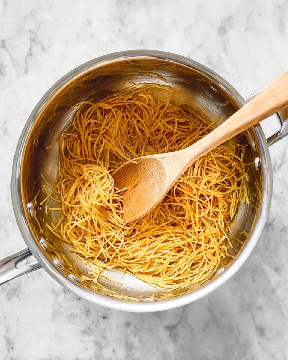 A close-up view of a shiny silver pot sitting on a white marbled surface with thin, golden brown toasted pasta strands inside, scattered unevenly as if stirring is in progress. A wooden spoon with a smooth texture lies diagonally across the pot, partially covered by the pasta strands. The pot's handles are visible but not the full sides, and there is a slight reflection on the pot's metal surface. photo taken with an iphone --ar 4:5 --v 7