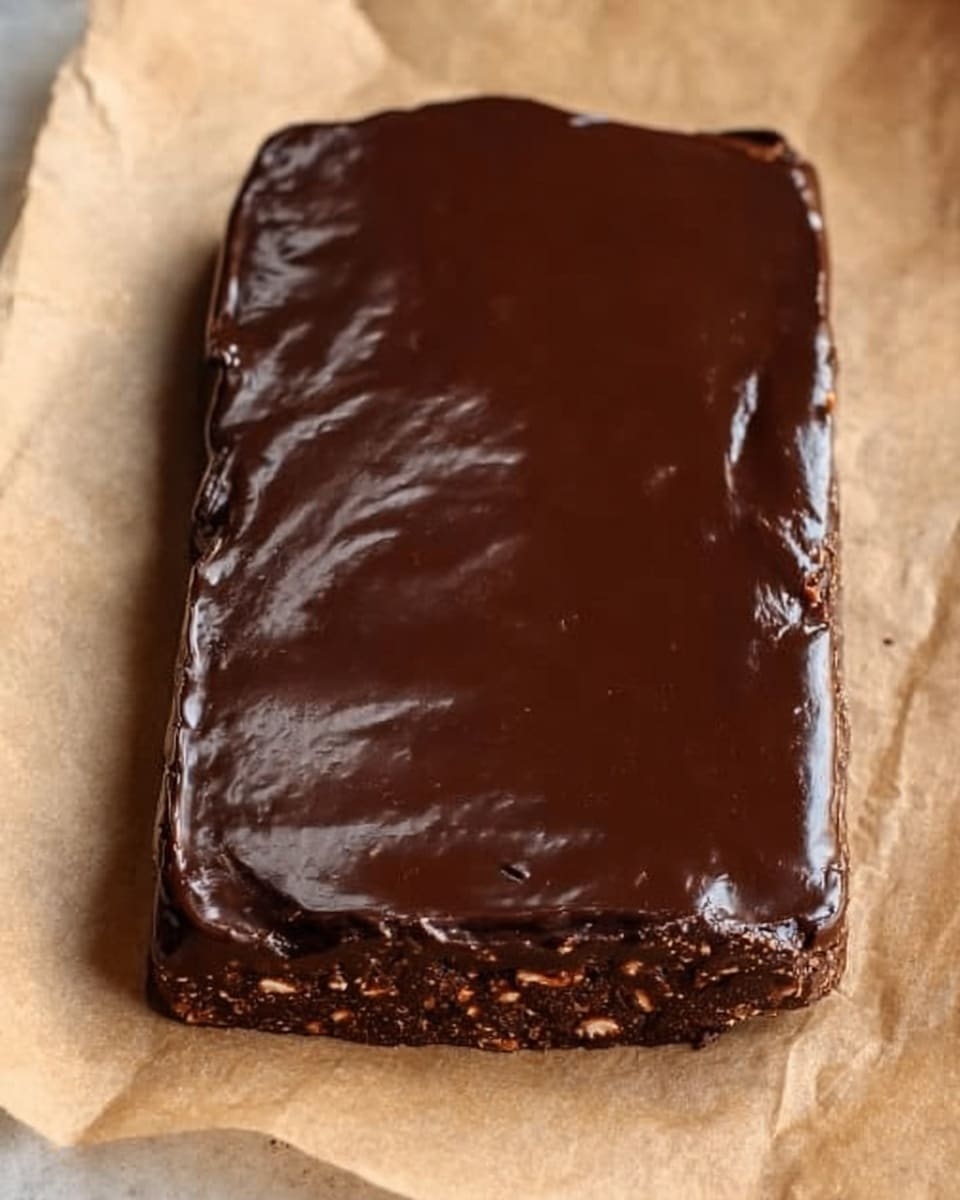 A rectangular bar with a thick bottom layer of dark brown, textured cookie or brownie base filled with small chunks, topped with a smooth, glossy layer of melted dark chocolate spread evenly, creating a rich and shiny surface. The entire bar is placed on a light brown parchment paper that contrasts with the dark colors of the bar. The edges of the chocolate layer slightly drip over the sides, showing a natural, handmade look. photo taken with an iphone --ar 4:5 --v 7