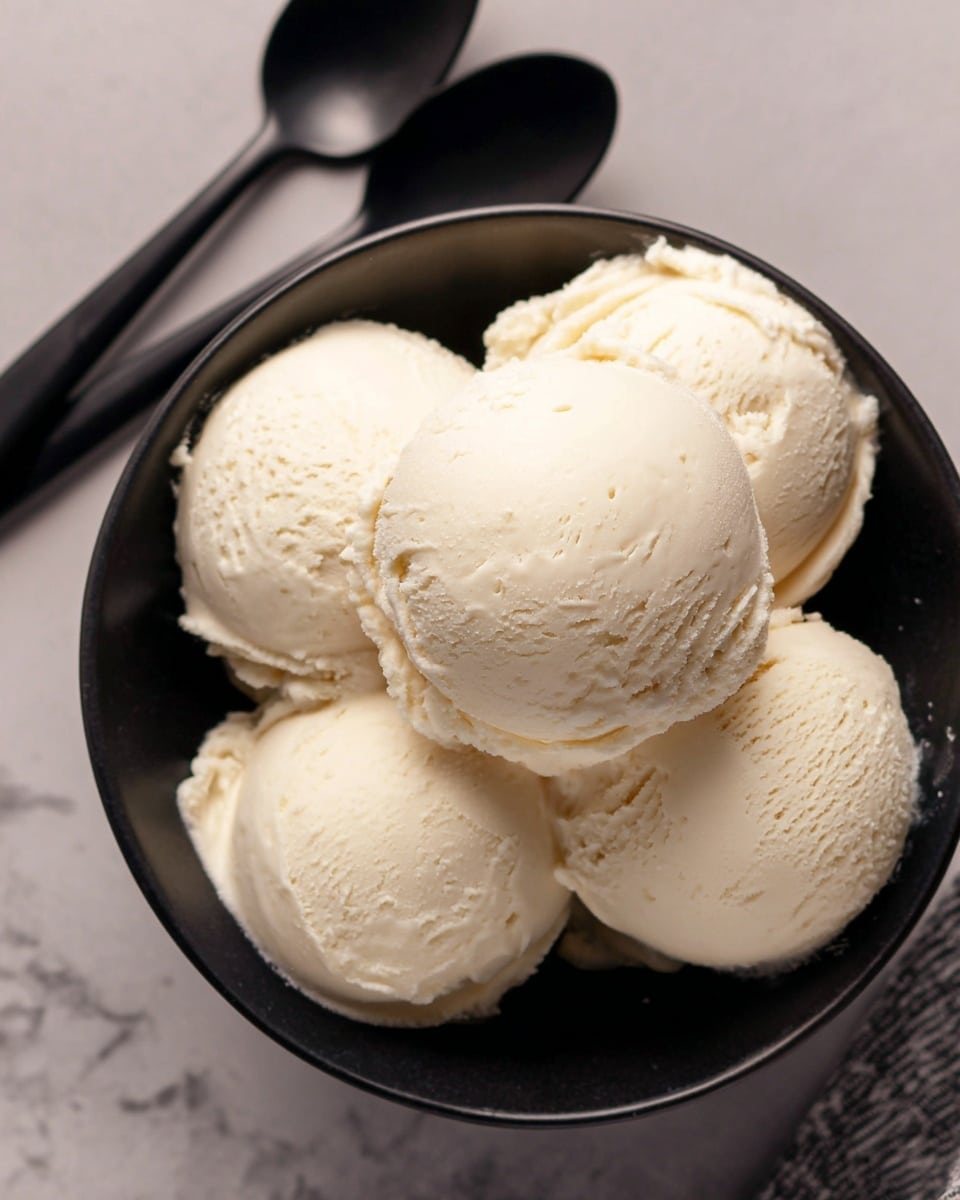 A bowl filled with five round scoops of smooth, creamy vanilla ice cream, all tightly packed together. The ice cream is pale off-white with a soft, slightly melting texture, giving a fresh and cool feel. The scoops sit inside a black bowl, set on a white marbled surface. In the background, there are two black spoons lying flat, one closer at the top left corner and the other at the bottom center. The image captures a top-down view, showing the round shapes and soft texture clearly. photo taken with an iphone --ar 4:5 --v 7