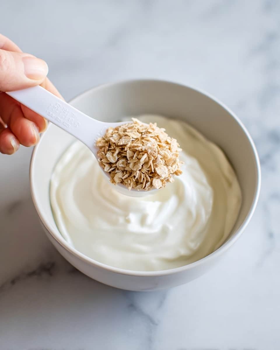 A close-up image shows a white bowl filled with thick white yogurt that has a smooth, creamy texture as the base layer. Above it, a woman's hand is holding a white measuring spoon filled with small, light brown dry flakes, positioned over the bowl as if about to add the flakes to the yogurt. The bowl sits on a white marbled surface, and the image has a clean, bright look. photo taken with an iphone --ar 4:5 --v 7