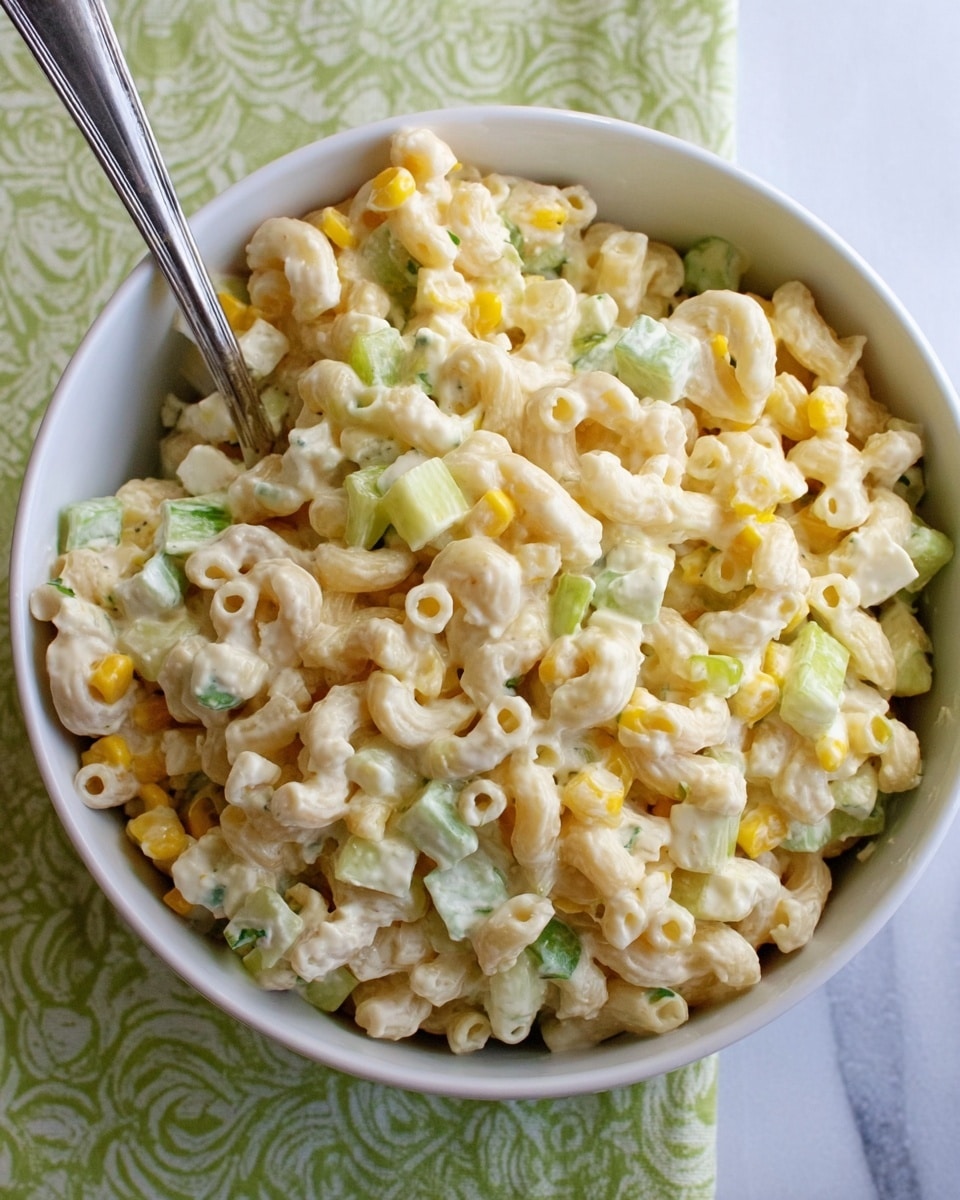 A white bowl filled with creamy pasta salad featuring small macaroni pieces lightly coated in a white dressing. Mixed throughout are pale yellow corn kernels, small cubes of white cheese, and light green celery pieces, creating a soft blend of colors and textures. A metal spoon rests inside the bowl, and the bowl is placed on a light green cloth with a pattern, all set on a white marbled surface. photo taken with an iphone --ar 4:5 --v 7