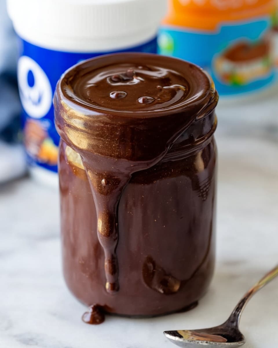 A clear glass dessert cup holds two scoops of creamy white ice cream with small dark specks mixed inside. The ice cream is topped with a thick layer of rich, dark chocolate sauce that drips slightly down the sides. In the background, out of focus, there is a dark brown jar with more chocolate sauce and a white container with blue accents. The whole scene is set on a white marbled surface. photo taken with an iphone --ar 4:5 --v 7