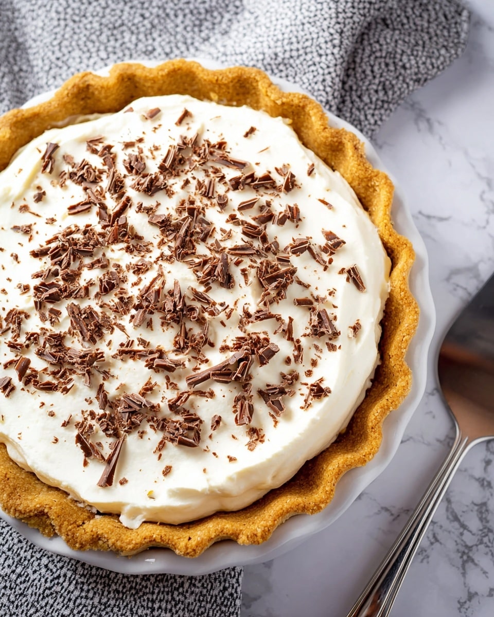 A close-up of a pie with a golden brown crust that has a crimped edge around the top. Inside the crust, there is a thick, smooth white layer of cream filling that covers the whole surface. On top of the cream layer, there are small, curled pieces of brown chocolate spread evenly across. The pie is on a white plate, placed on a white marbled surface with a silver pie server next to it. In the background, there is a folded gray and white patterned cloth. Photo taken with an iphone --ar 4:5 --v 7