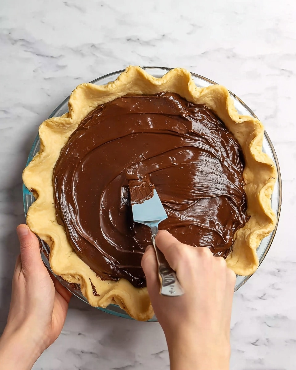 The image shows a pie with a golden, wavy crust on the outside, sitting on a white marbled surface. Inside the crust, there is a dark, shiny chocolate cream layer being spread smoothly with a spatula held by a woman's hand, while the other woman's hand holds the pie plate. The chocolate layer has a rich, smooth texture and covers the crust evenly. The pie is presented on a clear glass pie plate. Photo taken with an iphone --ar 4:5 --v 7