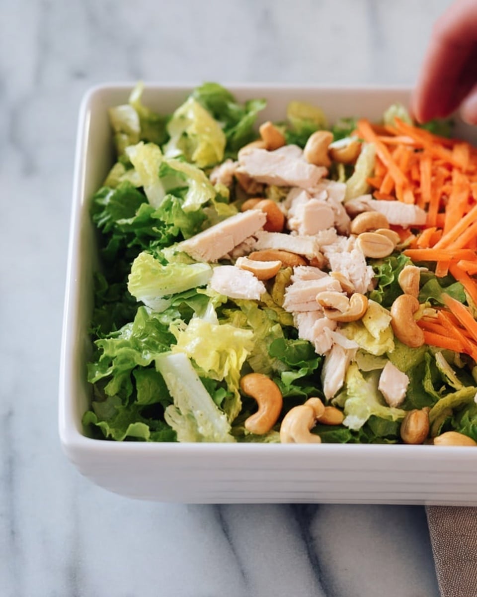 A white square bowl filled with a fresh salad that has three main layers: the base layer is a mix of bright green and pale yellow lettuce leaves with a crisp texture, the middle layer shows strips of orange carrot and light brown cashew nuts scattered evenly, and the top layer has small pieces of light beige cooked chicken. The bowl is placed on a white marbled surface, and a woman's hand is just touching the edge of the bowl. photo taken with an iphone --ar 4:5 --v 7