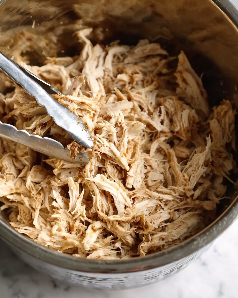 A close-up of a white rectangular dish filled with shredded chicken mixed with visible small red and black seasonings. In the background on a white marbled surface, there are three white round bowls: one with green leafy herbs, one with reddish sauce, and one empty or slightly blurred, with a halved lime next to them. The texture of the shredded chicken looks soft and tender with a slight shine. Photo taken with an iphone --ar 4:5 --v 7