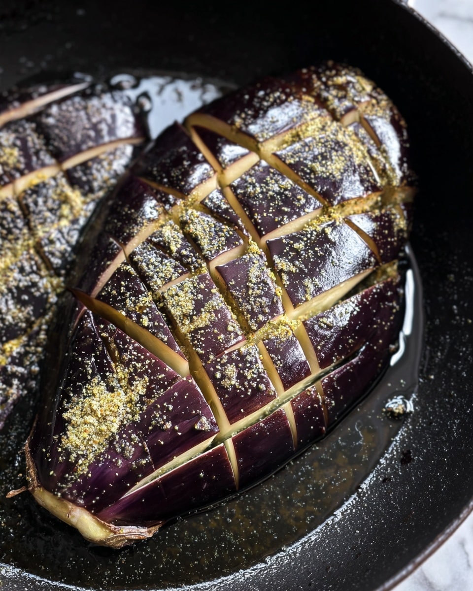 The image shows a deep purple eggplant with a shiny skin, sliced lightly in a crisscross pattern forming diamond shapes, lying flat in a black pan with oil. The surface of the eggplant is sprinkled with a greenish-yellow seasoning mix that looks grainy and slightly oily. The eggplant's pale inner flesh shows through the cuts, contrasting with the dark outer skin. The pan's black surface is dotted with small drops of oil and seasoning, giving a glossy texture. The scene is close-up, focusing on the eggplant's texture and seasoning details, on a white marbled surface. Photo taken with an iphone --ar 4:5 --v 7