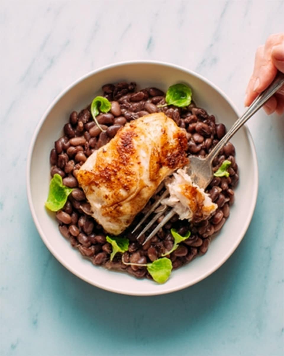 A white bowl filled with dark brown beans, arranged as the bottom layer with a soft, cooked texture. On top of the beans is a single piece of golden-brown cooked chicken, showing slightly crispy skin and tender meat underneath. Scattered small bright green leaves add a fresh touch around the chicken on the beans. A silver fork rests inside the bowl next to the chicken, and a woman's hand holds the fork. The bowl is set on a white marbled surface with a soft light that highlights the colors. Photo taken with an iphone --ar 4:5 --v 7