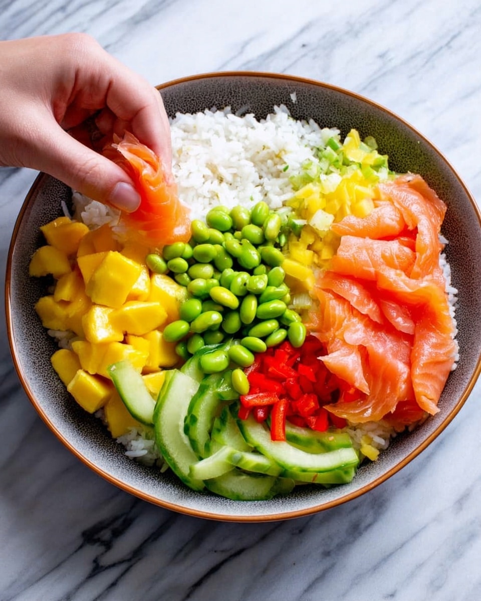 A round bowl with layers starting with white rice at the bottom, topped with neatly arranged sections of bright green edamame beans, yellow mango cubes, red bell pepper strips, and light green cucumber sticks. On one side, there are thin, folded slices of light pink smoked salmon being placed by a woman's hand. The bowl is on a white marbled surface. Photo taken with an iphone --ar 4:5 --v 7