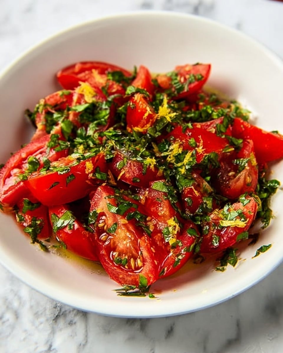 In a white bowl on a white marbled surface, there is a fresh salad made of bright red tomato wedges mixed with chopped green herbs. The tomatoes are glossy and slightly juicy, while the herbs add a textured green layer scattered evenly on top and throughout the salad. A light dressing with small bits of yellow zest or seasoning is visible, lightly coating the tomato pieces and herbs. The scene is bright and colorful, showing a fresh and simple dish. photo taken with an iphone --ar 4:5 --v 7