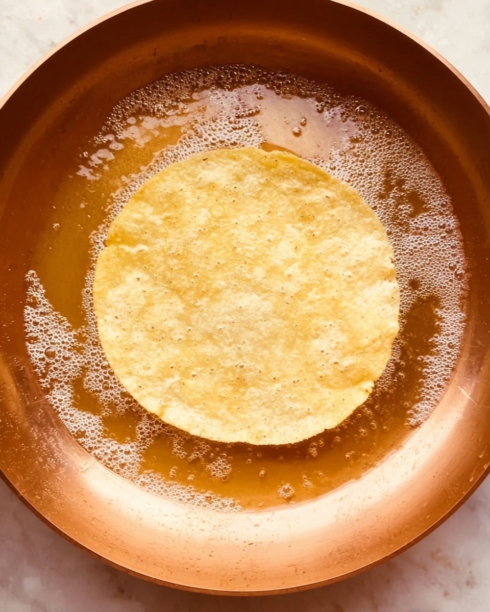 A single light yellow corn tortilla is frying in a copper-colored pan filled with bubbling oil, the edges of the tortilla slightly softening as it cooks. The tortilla is centered in the pan, which has a smooth texture with some oil reflections around the edges. The bubbles from the oil create a lively and warm feel, emphasizing the frying process. The background should be imagined as a white marbled surface. photo taken with an iphone --ar 4:5 --v 7