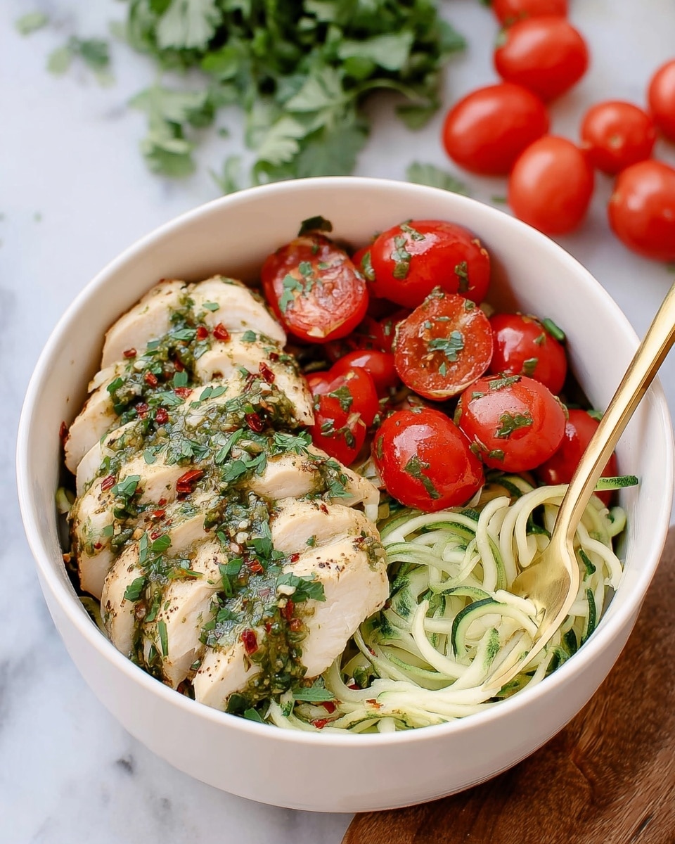 A white bowl sits on a white marbled surface filled with four distinct layers, starting with a base of light green spiralized zucchini noodles at the bottom right. On top and slightly left, there are five slices of lightly grilled white chicken, drizzled with a green herb sauce and sprinkled with chopped green herbs and red flakes. To the right of the chicken, a group of shiny, bright red cherry tomatoes with a few green herb bits scattered over them fills the remaining space. A gold fork is placed inside the bowl on the right side, and in the background, there are out-of-focus whole cherry tomatoes and some green herbs on the white marbled surface. Photo taken with an iphone --ar 4:5 --v 7