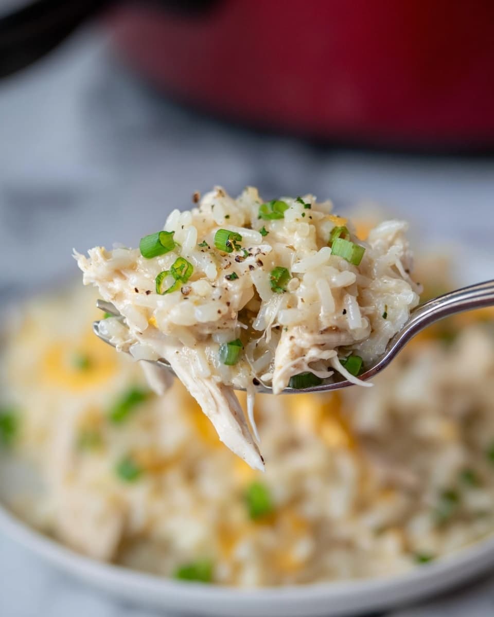 A close-up shows a fork holding a scoop of creamy rice dish with shredded chicken pieces mixed in. The rice is white and soft, coated with a light, creamy sauce that has a pale yellow tint. Small green chive pieces are sprinkled throughout, adding a fresh, bright green color. The details reveal specks of black pepper seasoning on the surface. The background is blurred but shows more of the rice dish on a white plate on a white marbled surface. Photo taken with an iphone --ar 4:5 --v 7