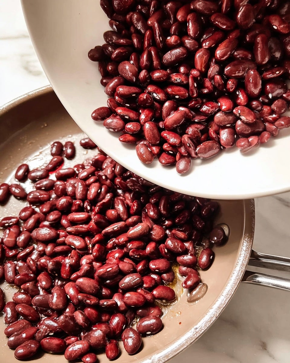 A close-up view of a white pan filled with dark red cooked beans. The pan is on a white marbled surface. A white plate is tilted, pouring a large amount of beans into the pan, causing many beans to scatter across the pan’s surface. The beans have a shiny, smooth texture and are packed closely together with some slight spacing around the edges. Photo taken with an iphone --ar 4:5 --v 7