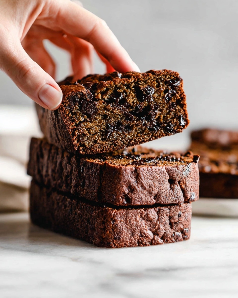 A close-up image shows a stack of three thick, rectangular slices of dark brown chocolate chip bread with a dense, moist texture and small chocolate chips embedded throughout. A woman's hand is picking up the top slice, highlighting its softness and rich color. In the background, more slices of the same bread are slightly out of focus, placed on a white marbled surface. The light is soft, bringing out the bread’s glossy, chocolatey details photo taken with an iphone --ar 4:5 --v 7