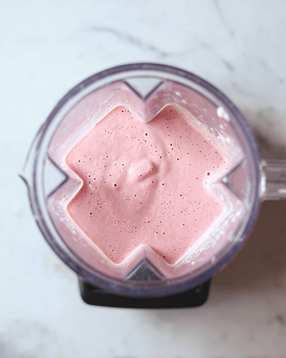 The image shows a clear blender jar from the top view, filled with a light pink, creamy mixture. The mixture has a smooth texture with small bubbles on its surface. The blender is placed on a white marbled surface, and the black base of the blender is slightly visible at the bottom of the image. Photo taken with an iphone --ar 4:5 --v 7