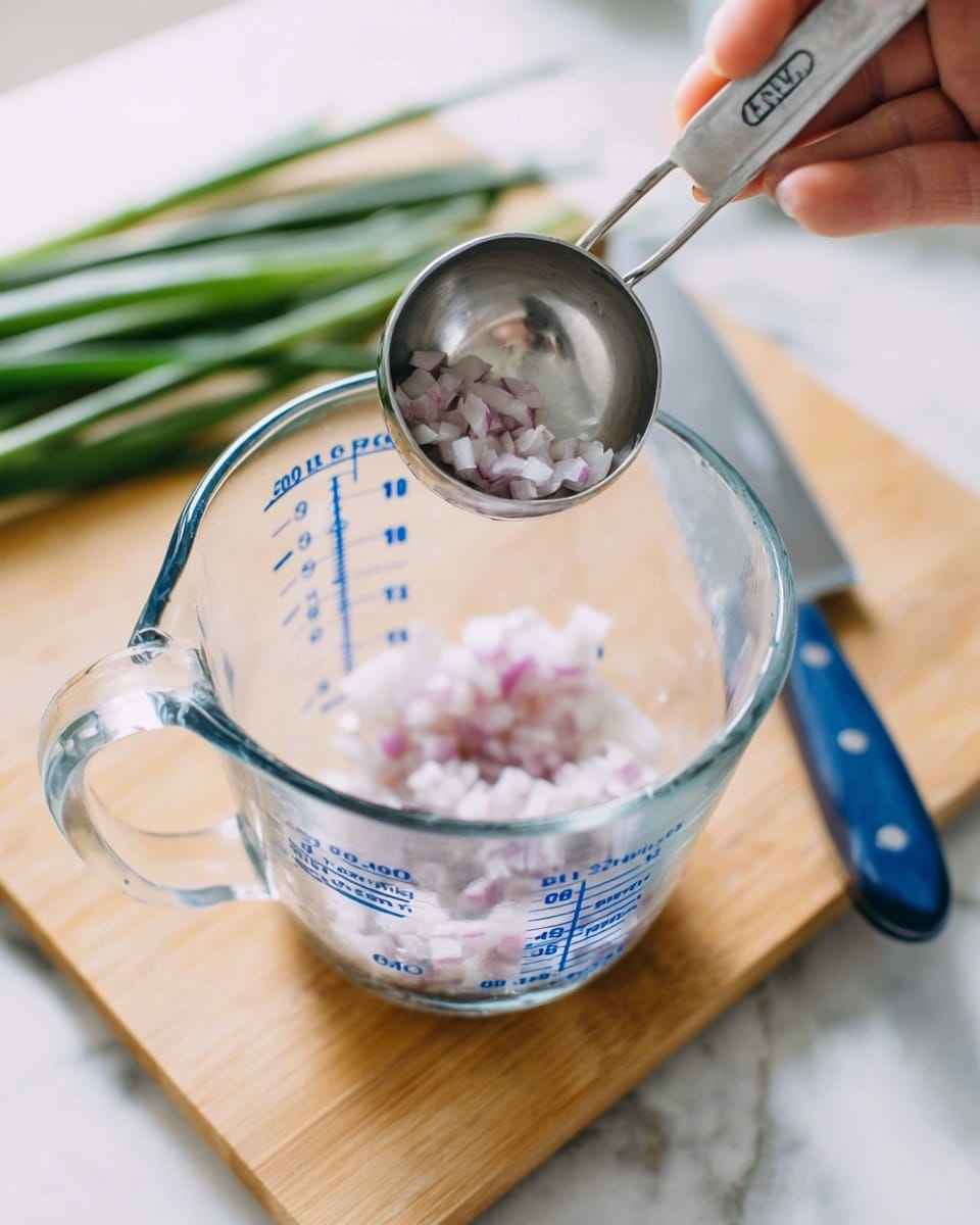 A close-up view of a clear glass measuring cup with blue numbers and letters, containing small chopped pale purple pieces, likely shallots or onions. Above the cup, a woman's hand holds a silver measuring spoon, poised as if to add an ingredient. The measuring cup sits on a light wooden board, beside a bunch of green onions on the white marbled surface. A blue-handled knife lies near the board, completing the kitchen scene. Photo taken with an iphone --ar 4:5 --v 7
