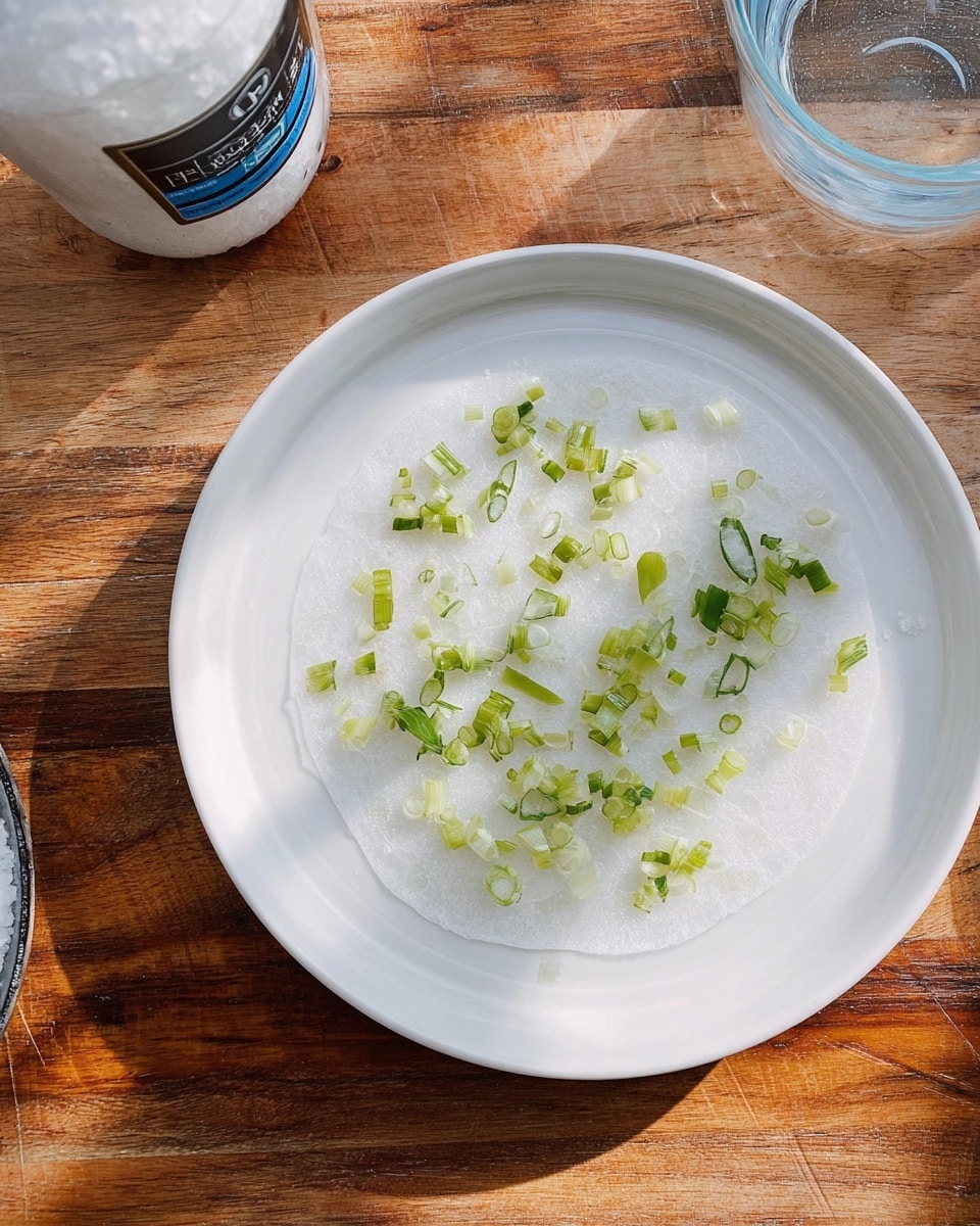 The image shows a white plate with one thin, round layer of translucent white rice paper placed flat on it. On top of the rice paper are small, scattered pieces of chopped green onions with varying shades of light and dark green. The plate sits on a wooden surface, but the background is changed to a white marbled texture. To the left of the plate, part of a white salt container with a blue label and a clear glass bowl are visible. photo taken with an iphone --ar 4:5 --v 7