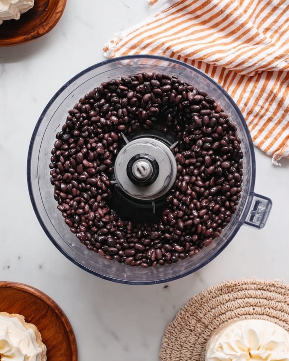 The image shows a clear food processor bowl filled with small black beans, evenly spread out in a single layer covering the entire inside surface. The food processor blade is visible in the center, surrounded by the dark, smooth beans with a slightly shiny texture. The food processor is placed on a white marbled surface. To the top right, there is a folded striped cloth with orange and white lines, and at the bottom right corner, there is a round white plate with a light brown knitted coaster under a small round cake decorated with a white flower on top. Photo taken with an iphone --ar 4:5 --v 7