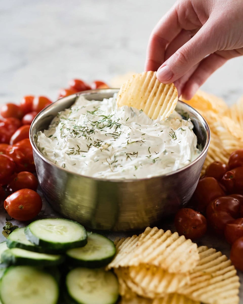 A close-up image shows a metal bowl filled with thick, white creamy dip sprinkled with green herbs on top. A woman's hand is dipping a ridged potato chip halfway into the dip. Surrounding the bowl are many small red grape tomatoes, green sliced cucumber pieces, and more ridged potato chips scattered around. All items sit on a white marbled surface. Photo taken with an iphone --ar 4:5 --v 7