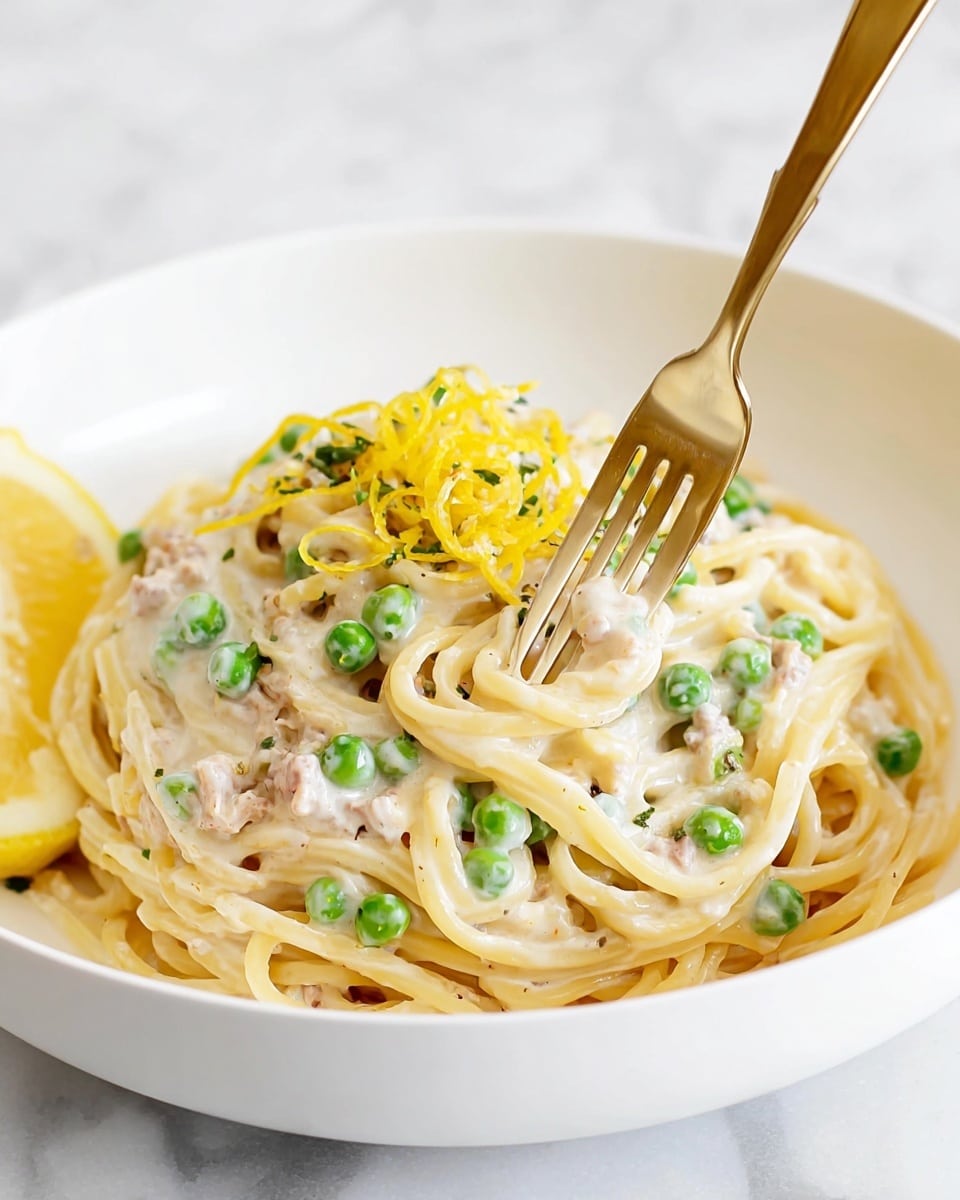 A white bowl filled with creamy spaghetti pasta showing two visible layers—the bottom layer is golden spaghetti noodles, while the top layer is thick white sauce mixed with green peas and small chunks of light brown meat. On top of the sauce is a small pile of thin yellow lemon zest curls and finely grated white cheese. A golden fork twirls some spaghetti and sauce near the center of the bowl, and a wedge of lemon rests against the side. The bowl is set on a white marbled surface. Photo taken with an iphone --ar 4:5 --v 7