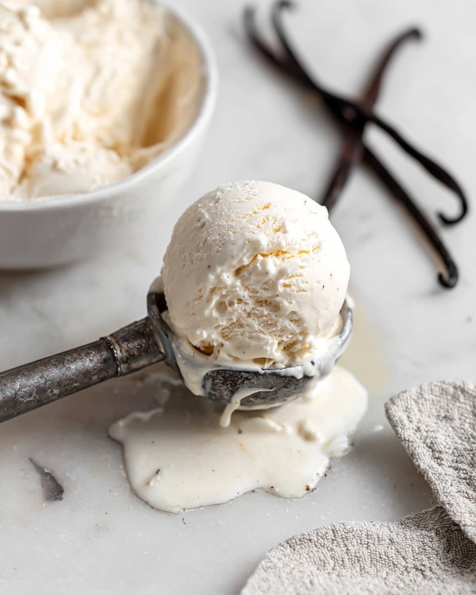 A scoop of pale creamy vanilla ice cream is held in an old metal ice cream scoop, showing its smooth, slightly melting texture with tiny vanilla bean specks throughout. The scoop is above a white marbled surface with some melted ice cream pooling and dripping down. Behind it, a white bowl filled with more pale vanilla ice cream is partially visible. Two dark vanilla pods lie on the white marbled surface to the right, and a light gray textured cloth is seen on the lower right corner. The photo taken with an iphone --ar 4:5 --v 7