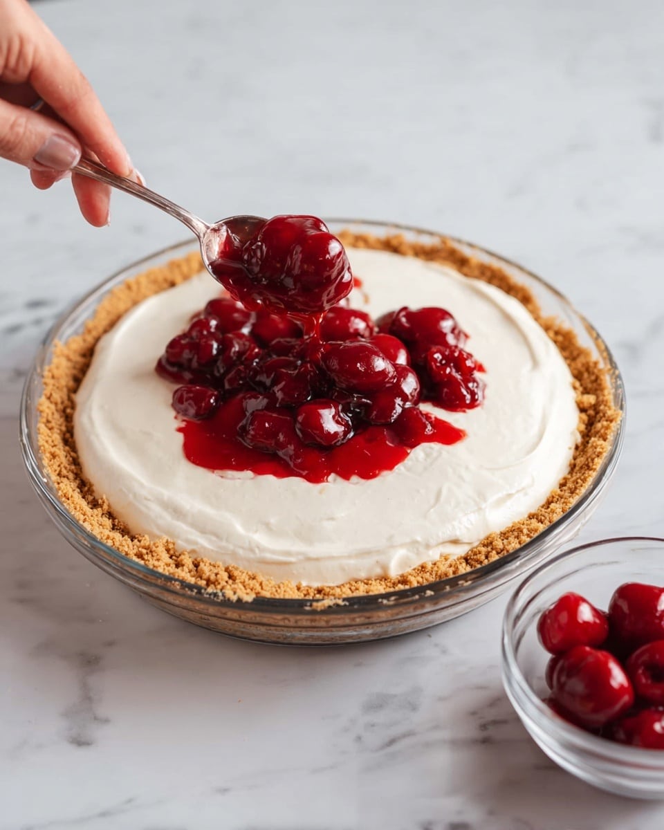 A pie with three layers is shown on a white marbled surface: the bottom crumb crust layer is light brown and crunchy, the middle layer is smooth and white creamy filling spread evenly, and the top layer is being added with bright red whole cherries in thick syrup, spooned from a silver spoon held by a woman's hand. The pie is in a clear glass dish, and a small clear bowl with more cherries is placed to the right. Photo taken with an iphone --ar 4:5 --v 7
