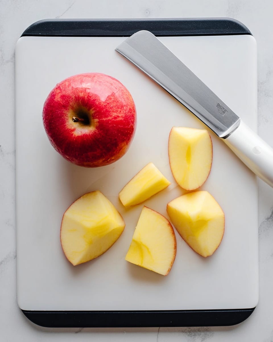 The image shows a white cutting board with a black edge on a white marbled surface. On the board, there is one whole red apple with a smooth, shiny skin. Next to the apple, there are six pieces of peeled apple arranged in two rows, showing a yellowish flesh with a smooth texture. A large, shiny kitchen knife with a white handle rests on the top right corner of the board, its blade positioned diagonally. Photo taken with an iphone --ar 4:5 --v 7