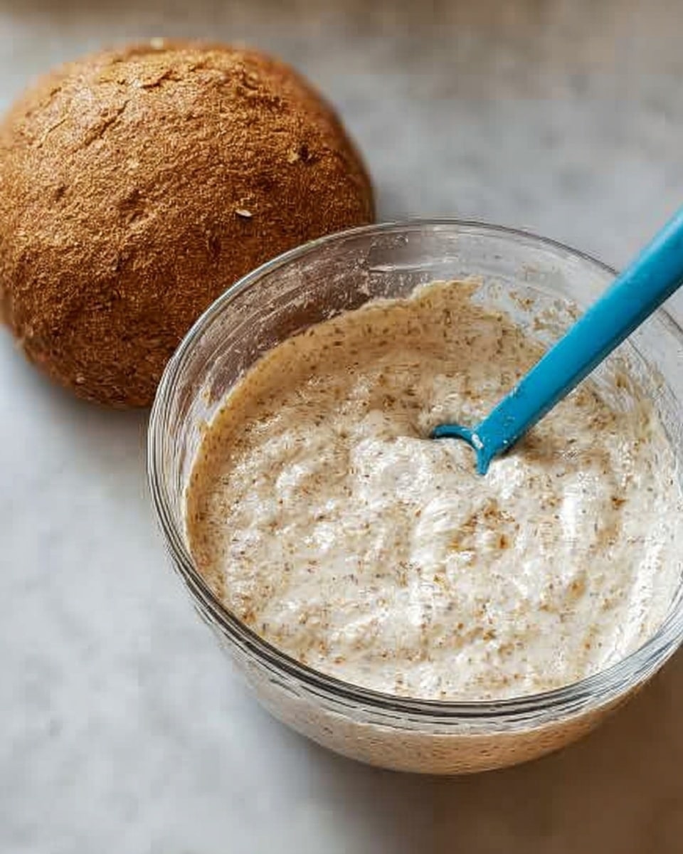The image shows a clear glass bowl filled with a thick, creamy mixture that is light beige with specks of darker grains throughout, giving it a textured look. A bright blue spoon is placed inside the bowl, slightly submerged in the mixture. Next to the bowl, there is a round, brown bread roll with a rough, crackled crust. Both the bowl and bread rest on a white marbled surface. photo taken with an iphone --ar 4:5 --v 7