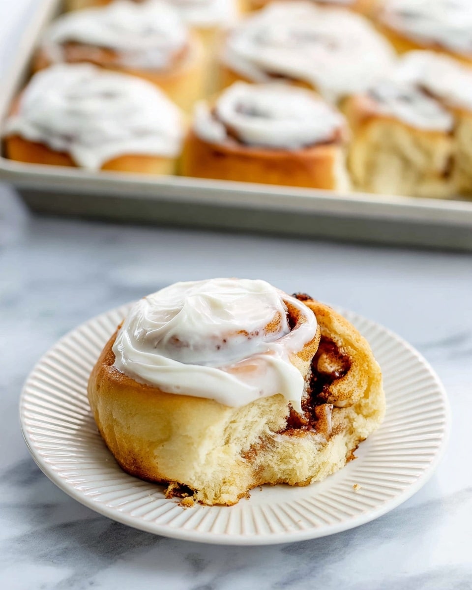 A single cinnamon roll sits on a white plate with ridged edges, its soft golden-brown dough partially torn to reveal a dark brown cinnamon swirl inside. On top, a thick layer of smooth white cream cheese frosting is spread unevenly, showing some swirl patterns. In the background, more cinnamon rolls, each topped with the same white frosting, rest closely together on a baking tray. The surface beneath everything is a white marbled texture. photo taken with an iphone --ar 4:5 --v 7
