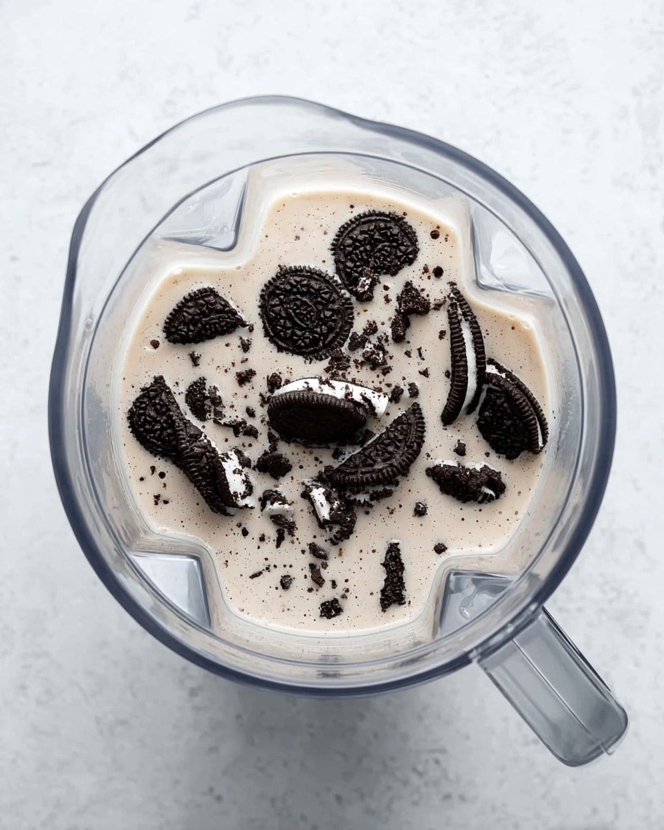 A clear blender jar sits on a white marbled surface, filled with a light beige liquid topped with many small and large pieces of black chocolate sandwich cookies with white cream filling, scattered unevenly across the liquid's surface. The blender jar is clean and transparent with a sturdy handle on the right side, showing clear lines and details inside. The image is bright and simple, focusing on the mix inside the blender. photo taken with an iphone --ar 4:5 --v 7