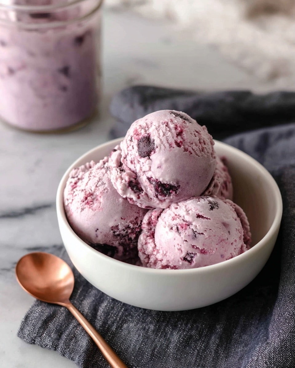 A white bowl holds three scoops of light purple and pale pink ice cream with small dark berry bits mixed inside, giving a creamy and slightly rough texture. The bowl is placed on a dark gray cloth over a white marbled surface. Next to the bowl is a plain copper spoon. In the background, there is a glass jar filled with the same purple and pink ice cream. Photo taken with an iphone --ar 4:5 --v 7