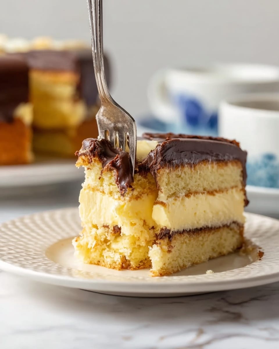 A close-up of a cake slice on a white plate with a textured edge, showing three layers: a light golden brown cake layer at the bottom, a thick creamy yellow middle layer with a soft texture, and a smooth dark chocolate layer on top that looks shiny. A silver fork is cutting into the cake with some of the creamy middle and chocolate topping on the fork. In the blurred background, there is another white plate with the rest of the cake and a white cup with a blue pattern, all set on a white marbled surface. Photo taken with an iphone --ar 4:5 --v 7