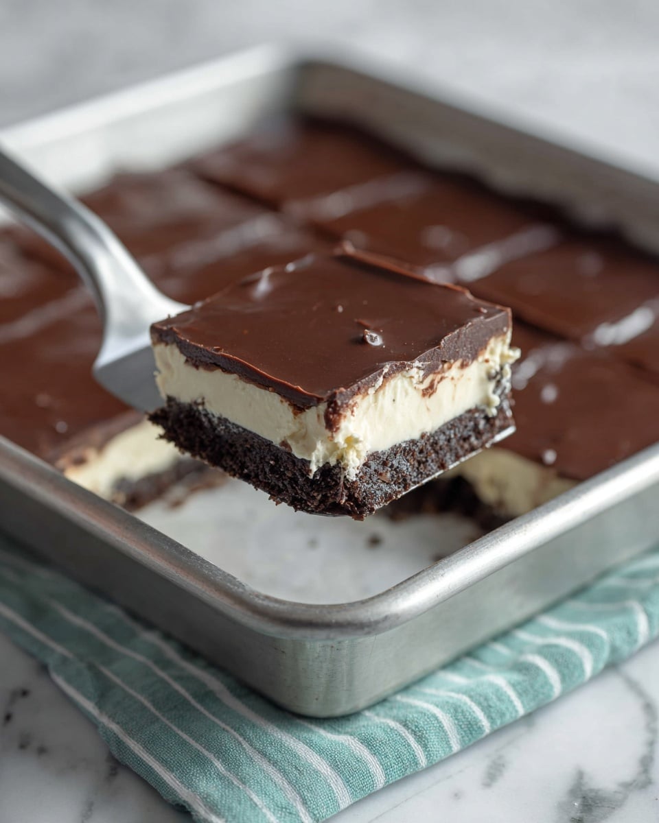 A square piece of dessert with three layers is being lifted by a metal spatula above a metal baking tray. The bottom layer is dark brown and crumbly, the middle layer is thick and creamy white, and the top layer is smooth and glossy dark brown chocolate. The baking tray is on a teal and white striped cloth, sitting on a white marbled surface. The piece above the tray shows neat edges and a shiny chocolate top with small marks. Photo taken with an iphone --ar 4:5 --v 7