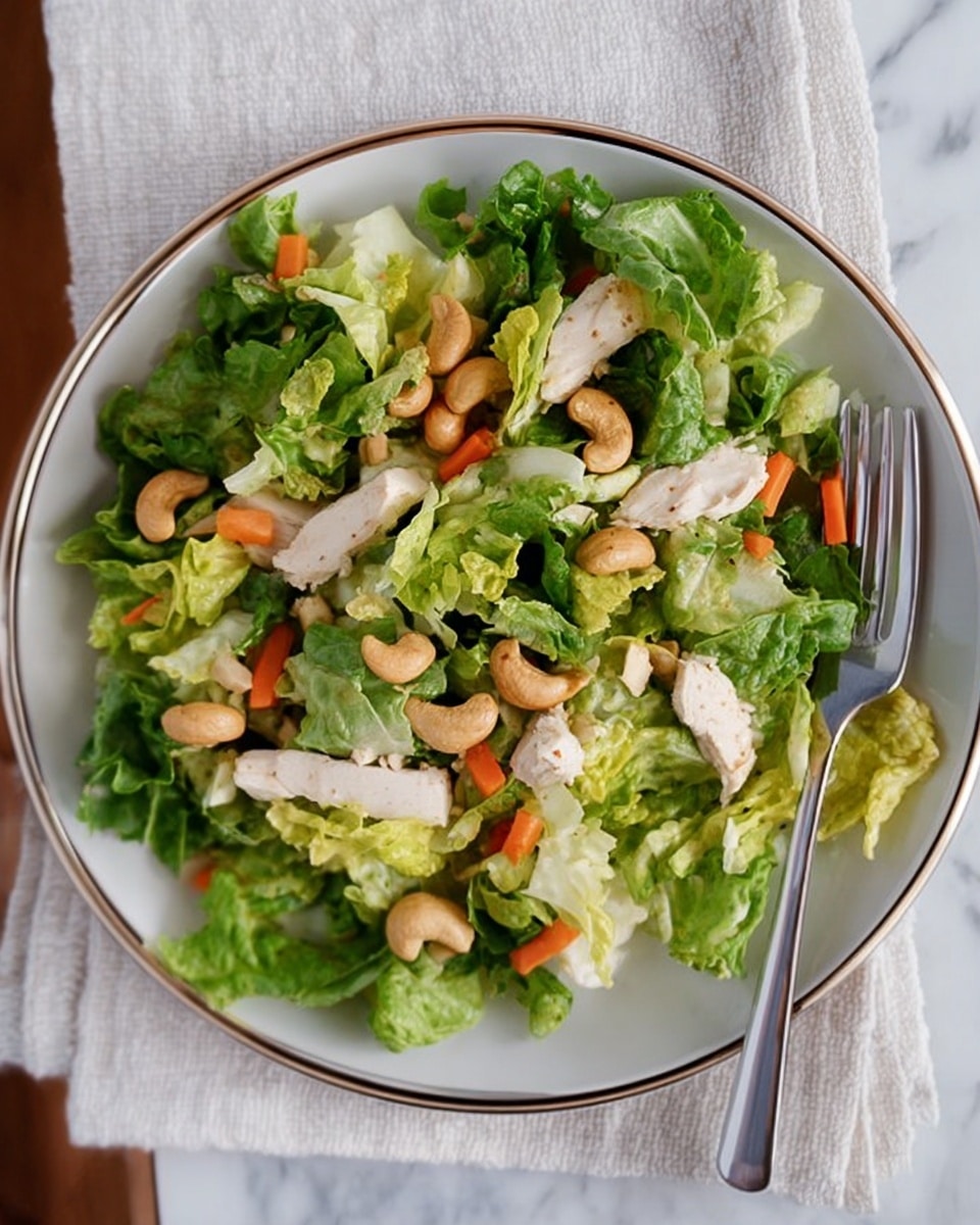 A fresh salad served on a white plate with a metallic rim, resting on a white cloth over a white marbled surface. The salad has three main layers: the base layer of bright green chopped lettuce with some light yellow leafy edges, a middle layer of diced white chicken pieces, and scattered orange carrot sticks. The top layer features whole light brown cashew nuts spread evenly across. A fork is placed on the right side of the plate among the salad greens. The overall look is fresh, colorful, and inviting. photo taken with an iphone --ar 4:5 --v 7