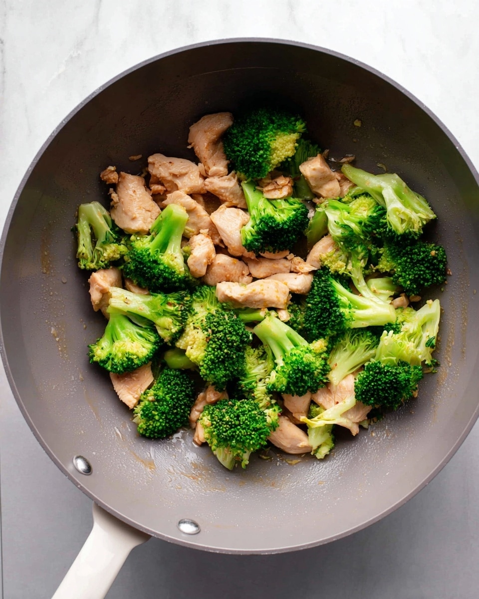 Inside a grey wok, there is a mix of light brown cooked chicken pieces and bright green broccoli florets scattered together, showing the contrast between the soft, pale chicken and the fresh, textured broccoli. The wok sits on a white marbled surface, adding a clean and fresh look to the image. The chicken pieces are uneven but tender-looking, while the broccoli pieces have a crisp, slightly shiny surface as if just cooked. Photo taken with an iphone --ar 4:5 --v 7