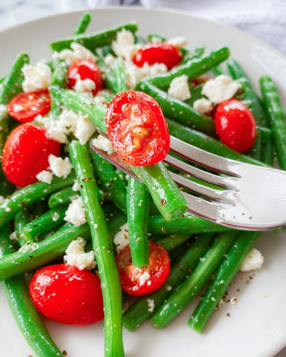 A close-up view of a fresh green bean salad on a white plate with a white marbled surface below. The salad has one layer of bright green beans that are crisp and smooth, mixed with halved red cherry tomatoes scattered throughout. Small white crumbles of cheese are sprinkled evenly on top, adding a soft texture contrast. A shiny silver fork presses into one of the cherry tomato halves, showing its juicy inside. The overall colors are bright green, red, and white, with a few black pepper specks adding texture. Photo taken with an iphone --ar 4:5 --v 7