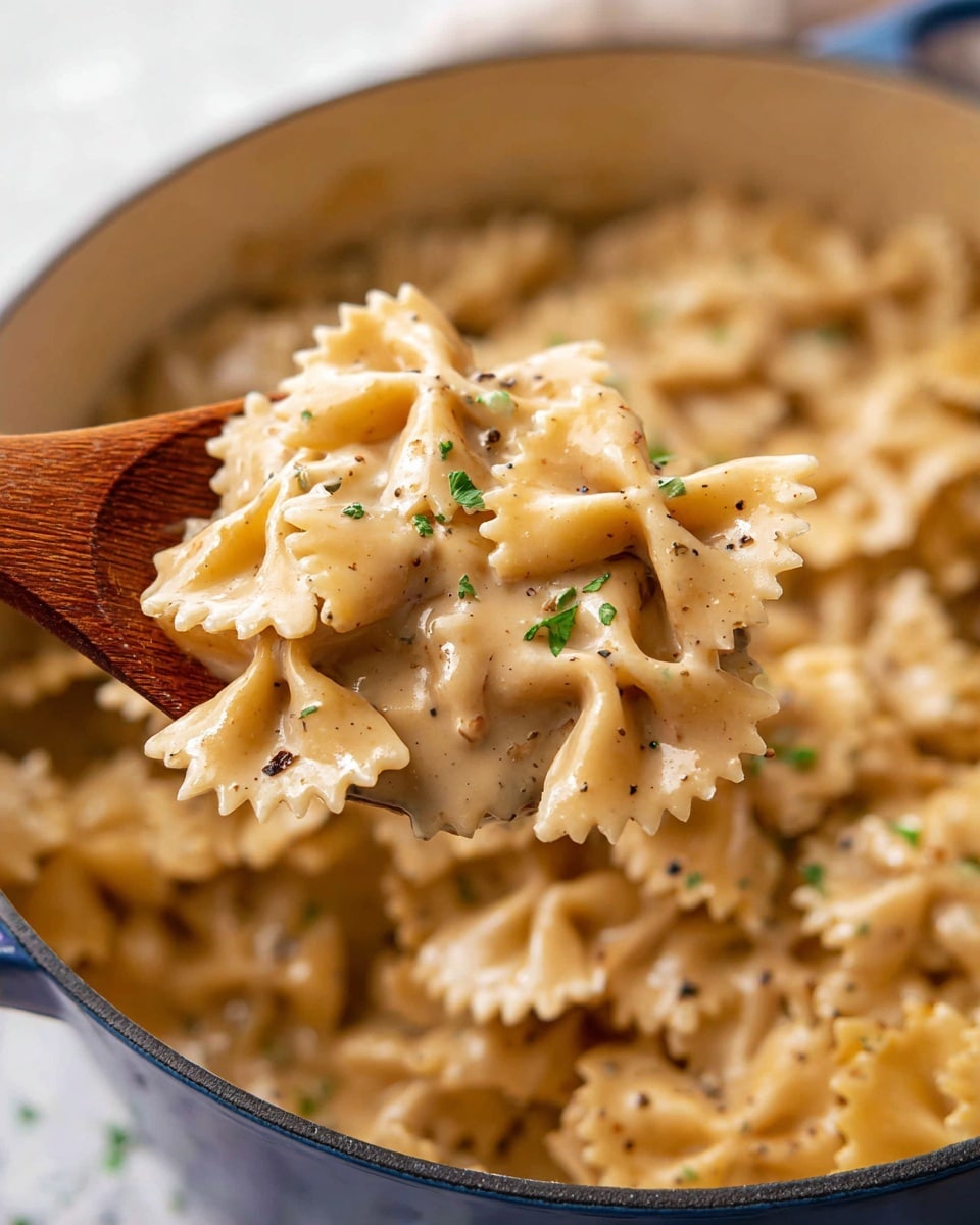 A close-up of creamy bowtie pasta held by a wooden spoon over a white pot filled with the same pasta. The bowtie noodles are coated in a smooth, light beige sauce, with flecks of black pepper and small green herbs sprinkled on top. The sauce looks rich and slightly creamy, covering each pasta piece evenly. The background surface has a white marbled texture and the overall setting feels warm and inviting. Photo taken with an iphone --ar 4:5 --v 7