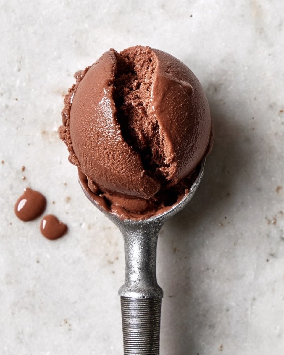 A single scoop of dense, smooth chocolate ice cream with a rich dark brown color sits firmly on a shiny metal ice cream scooper with a textured handle. The scoop has a slight fold showing creaminess and a glossy spot reflecting light. A few small drops of melted chocolate ice cream are near the bottom left on a white marbled surface photo taken with an iphone --ar 4:5 --v 7